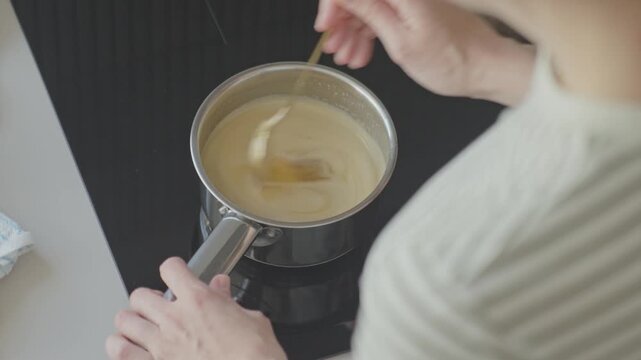 Woman stirring creamy sauce in saucepan on induction cooktop. Concept of home cooking and dessert preparation