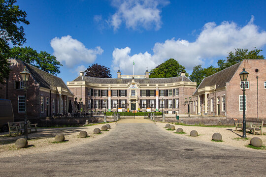 View of Castle Groeneveld historic estate with its grand brick facade, side wings, and gravel driveway under a blue sky with white clouds in Baarn, Utrecht, Netherlands.