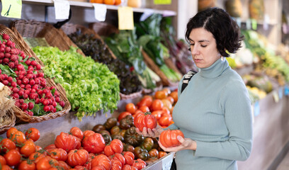 Middle-aged woman buys ripe red tomatoes in a supermarket. European woman stands near a vegetable counter and holds tomatoes in her hands, examines them and chooses them to cook dinner © JackF