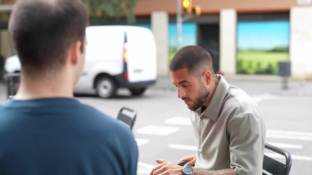 Two men converse outdoors. One listens while the other speaks animatedly. Parked van and building form the backdrop. The scene captures an informal, engaging discussion