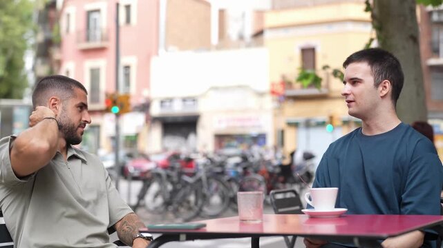 Two men converse at a cafe table. They smile, engaged, with coffee and phones nearby. Background shows bicycles and buildings. Urban setting, casual, relaxed interaction. Ideal for social, friendship