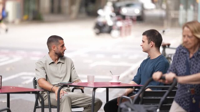 Two men chat at a cafe table. Woman rides a bike past them. City streets blur behind them. Natural, calm conversation in public space. Ideal for urban lifestyle or everyday moments
