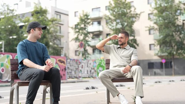 Two men converse on a public bench. One man gestures while speaking, the other listens attentively. Trees and graffiti frame their urban setting. Natural light enhances the casual, relaxed atmosphere