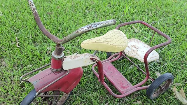 Rusty red vintage tricycle on green grass during a downward tracking shot, with faded paint, worn wheels, and cream seats in an outdoor setting.