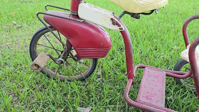 Close view of a vintage red tricycle with rusty metal parts and cream seat on green grass during an upward tracking shot outdoors.