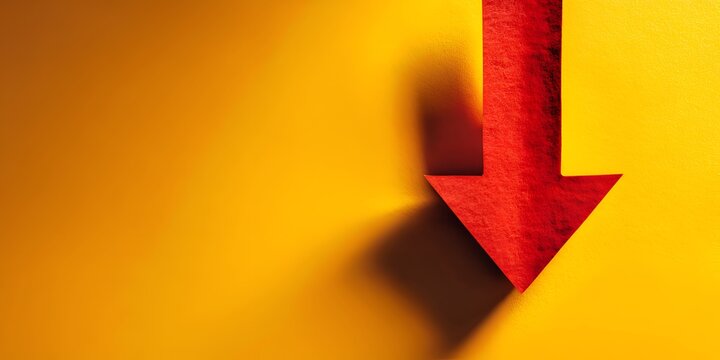 A textured red arrow dramatically points downwards on a vibrant yellow surface casting a stark shadow indicating decline or negative trend.