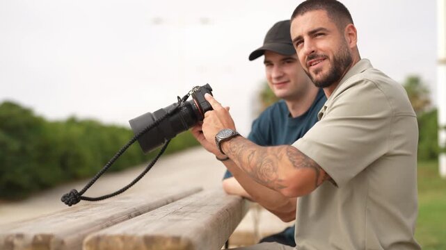 Man holds camera, leans on bench. Tattooed arm shows confident gesture. Second man watches, smiling behind. Outdoor setting suggests casual photography session. Natural light enhances candid moment
