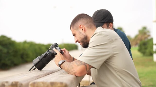Man holds camera, focused on scene ahead. He leans on bench, eyes fixed on subject. Second man stands behind, observing quietly. Outdoor setting, soft light, calm atmosphere