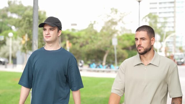 Two men walk side by side through a park. One wears a cap and dark t-shirt. The other sports a polo and beard. They face forward, calm, unbothered. Urban trees line the path behind them