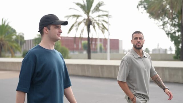 Two men walk side by side, casually conversing. One wears a cap, the other sports a short beard and visible tattoo. They stroll past palm trees and a concrete barrier