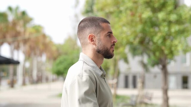 Man walks calmly through urban park. He wears light-colored collared shirt. Background features trees and modern buildings. His expression shows quiet focus or thought. Ideal for lifestyle, urban