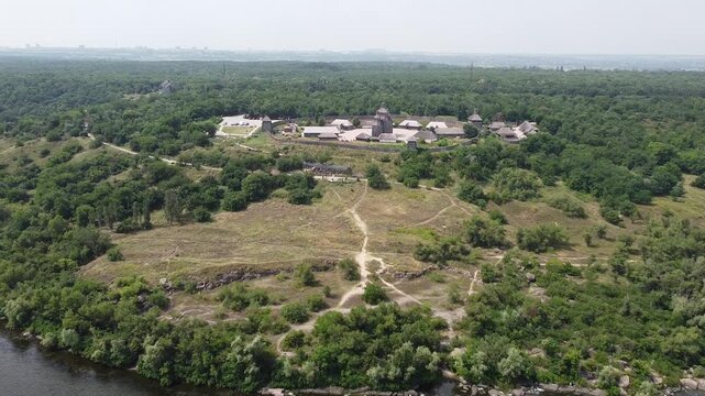 Horizontal drone footage of Khortytsia Island in Zaporizhzhia, Ukraine, featuring a historic wooden settlement surrounded by a wooden fence, lush green forests, and the Dnipro River