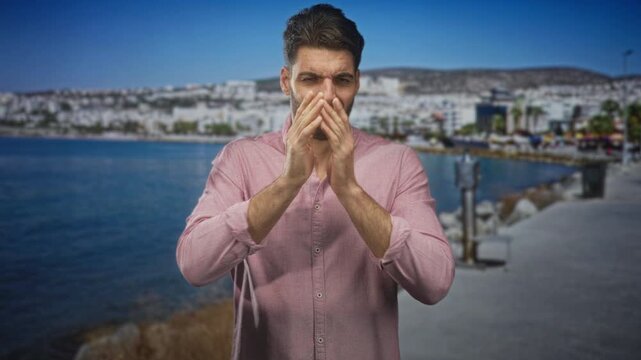 Man with beard covering his face with hands, wearing a pink button shirt, standing on a seaside street by a harbor pier; embarrassment vulnerability reflection.