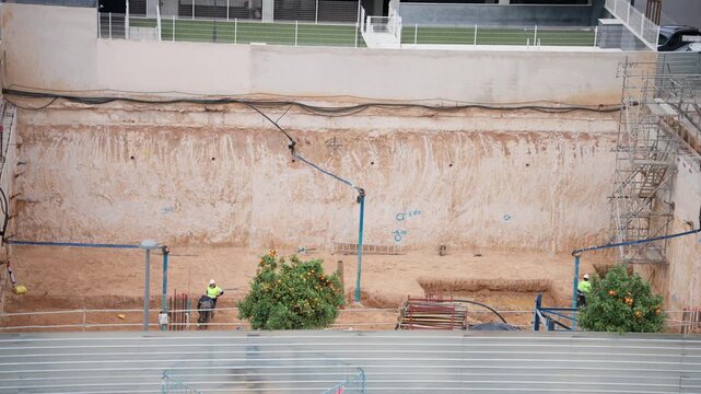 Two workers in yellow safety vests stand inside a deep excavation pit next to reinforcement rods, surrounded by orange trees on sandy ground with a concrete retaining wall