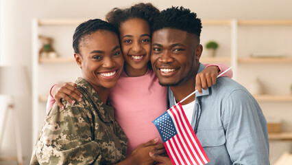 A military service member reunites with family in a warm indoor space. They are smiling and holding a small American flag. The daughter stands between her parents, all look happy. © Prostock-studio