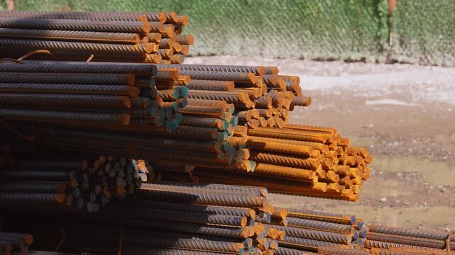 Large bundles of rusty steel reinforcing bars stacked outdoors at a building construction site, with a green mesh fence in the background and a gentle breeze rustling a piece of paper