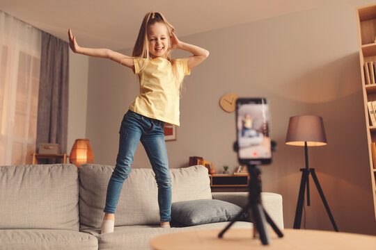 A young girl enjoys dancing on a couch in her living room. She performs playful moves while recording a video with a smartphone set on a tripod, showcasing her energy and joy.