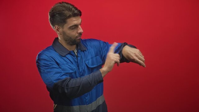 Man in blue work overalls points index finger and taps wristwatch in studio red backdrop; professional punctuality focus.