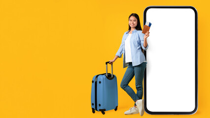 A woman smiles while holding a phone and travel documents. She stands next to a blue suitcase against a bright yellow background. This scene shows excitement for an upcoming journey. © Prostock-studio