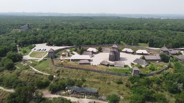 Horizontal drone footage of Khortytsia Island in Zaporizhzhia, Ukraine, showing a historic wooden Cossack settlement enclosed by a wooden fence, surrounded by lush green forests and the Dnipro River