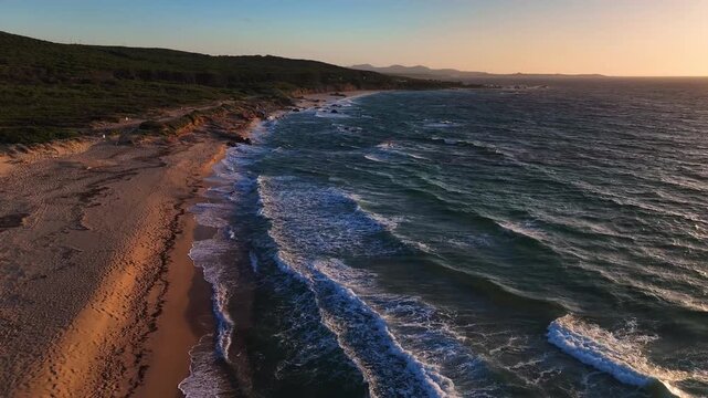 Aerial view of the sandy beach at Costa Paradiso with waves crashing on the shore and green hills under a golden sunset light Costa Paradiso, Sardegna, Italy.