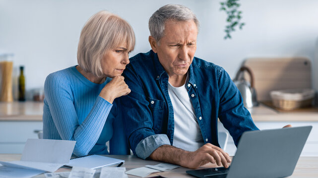 Two older adults are seated at a table filled with papers, looking at a laptop. Their expressions are focused as they review financial documents together. Sunlight fills the room.