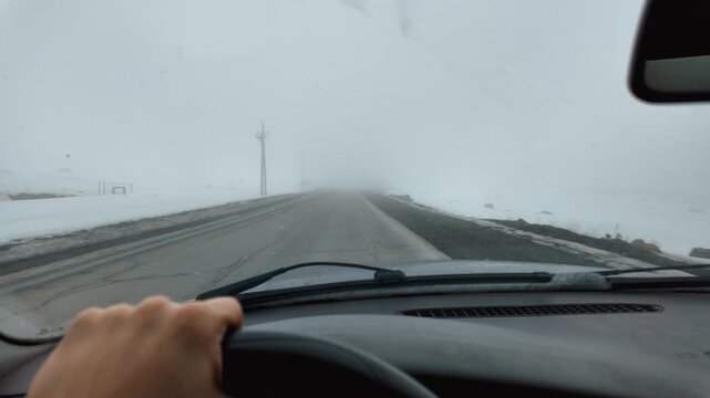 Point of view from a car driving on a dangerous, empty road during a foggy winter day, with a hand firmly on the steering wheel navigating the poor visibility and snowy landscape
