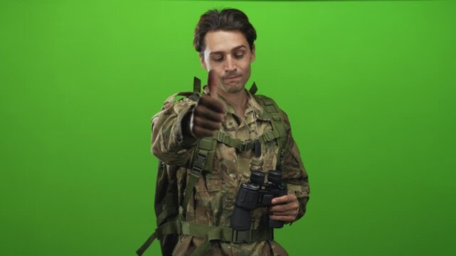 Soldier in camouflage uniform wearing backpack and dog tags, holding binoculars and giving thumbs up to camera in studio; approval duty morale.