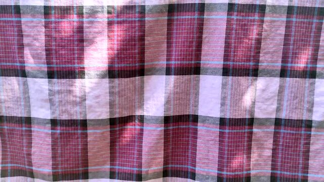 Traditional plaid pattern fabric sarung waving in the wind with natural sunlight and shadows. Red and black checkered textile background. Close up of soft material texture moving outdoors