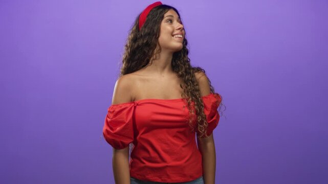 Hispanic woman smiling with bare shoulder visible, wearing red off shoulder top and headband, looking sideways against a purple studio wall; youthful joy.