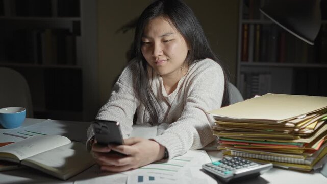 Woman holding smartphone and resting chin on hand at cluttered study desk with open books, stacked folders, calculator and blue coffee cup in building; study fatigue.