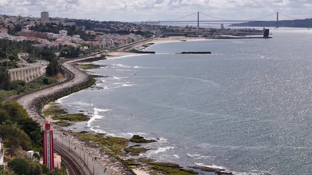 Aerial view of Passeio Maritimo de Alges with the red lighthouse, railway tracks, and the 25 de Abril Bridge in the distance in Cruz Quebrada, Lisboa, Portugal.