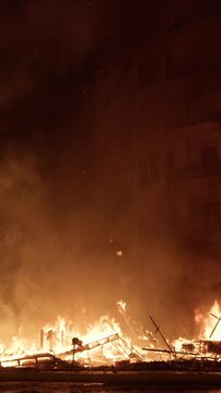 Vertical frame of smoldering falla debris after La Crema burning in Valencia, thick smoke rising against dark orange sky with glowing ember particles drifting upward into darkness