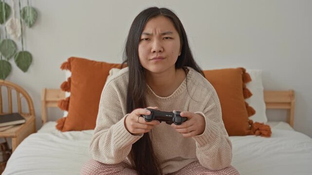 Woman holding a game controller, hands gripping the controller on a bed in a bedroom; focused concentration.