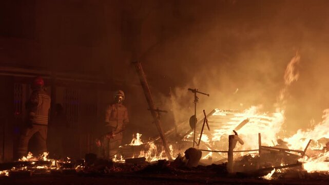 Firefighter in protective gear and white helmet silhouetted against burning collapsed falla debris during Las Fallas La Crema in Valencia, second firefighter visible amid smoke and orange flames