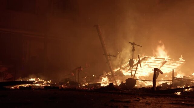 Single firefighter with white helmet and protective gear stands watching burning wooden remains of a collapsed falla during Las Fallas La Crema celebration in Valencia at night