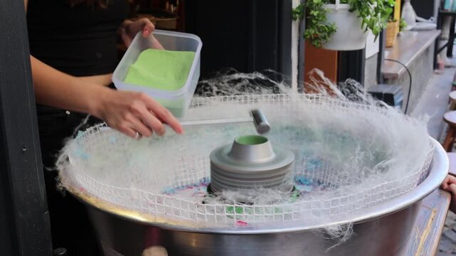 Medium close video of vendor making green cotton candy with spinning machine and sugar powder, showing street dessert preparation, confectionery craft, snack service, festival food, and sweet treat.
