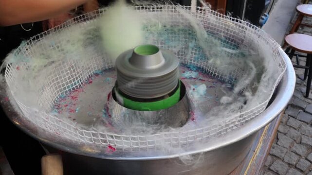 Close view video of vendor wrapping cotton candy on a stick and handing it to customer near the spinning machine, showing street snack preparation, serving process, confectionery trade, retail.