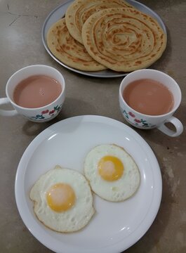 Two fried eggs on a white plate, beside floral cups of tea, breakfast time