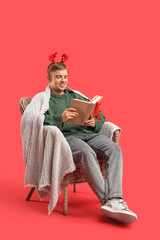 Naklejka premium Young man reading book while sitting in armchair on red background. Christmas celebration