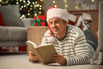 Naklejka premium Young man in Santa hat reading book at home on Christmas eve