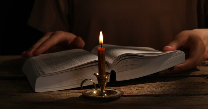 Woman reading Bible at wooden table with burning candle, closeup