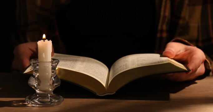 Woman reading Bible at wooden table with burning candle, closeup. Casting light and shadow