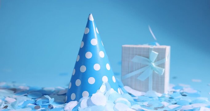 Party hat and gift box under falling beautiful confetti on light blue background, closeup