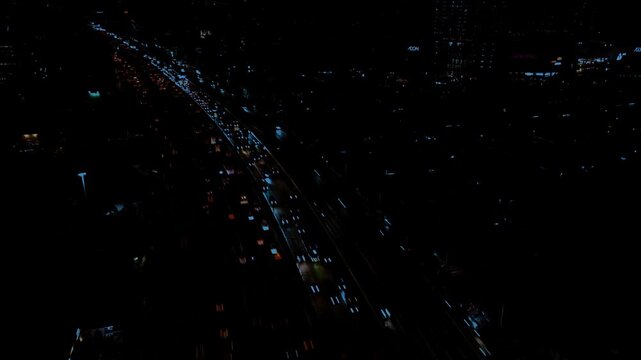 Static aerial shot of glowing city lights and moving vehicles on a multi-lane expressway during the dark night hours in blue ish color graded.