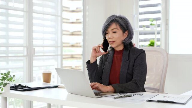 Senior businesswoman having call on smartphone with business partner.  Entrepreneur talking by mobile cellphone while working on laptop computer in office.