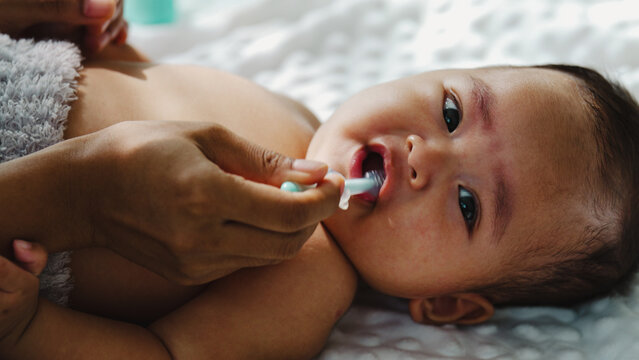 Soft light bathes a baby's calm face. Gentle hand brushes the child's teeth with care. The baby gazes upward, eyes wide and trusting. Tiny gums and a curious mouth frame the moment