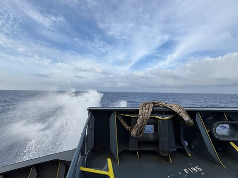View from the bow of a moving ship cutting through ocean waves, with strong water spray on both sides, dramatic cloudy sky, and visible deck structure emphasizing motion, power