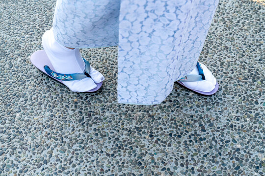 Close-up of person wearing traditional Japanese white tabi socks and purple zori sandals on a pebble path. Light blue floral kimono details shown from high angle. Cultural fashion and travel concept.