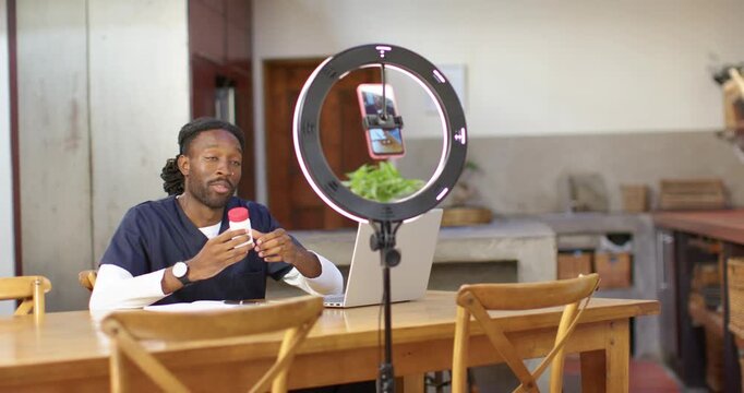 African American man in scrubs mounting phone in ringlight in kitchen filming demo with red bottle